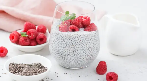 Pudding chia z malinami i miętą w szklance, obok miseczki z nasionami chia i malinami.