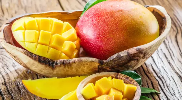 Dwa soczyste mango, jedno w całości, drugie pokrojone w kostkę, w drewnianych miseczkach. Idealne na zdrową przekąskę, mango kcal to ok. 60 na 100g.