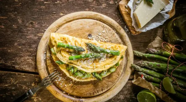 Pyszny omlet ze szparagami, posypany parmezanem, czeka na widelcu. Obok świeże szparagi i kawałek sera.