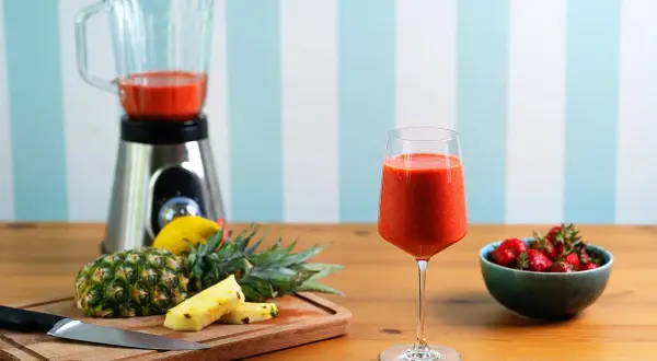 Pyszny koktajl owocowy w kieliszku, obok ananas, truskawki i blender.
