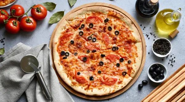 Pyszna pizza bez glutenu z pomidorami i oliwkami, gotowa do pokrojenia. Obok świeże pomidory, oliwa i grissini.