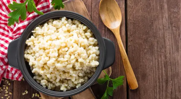 Kasza bulgur w czarnej misce, obok drewniana łyżka i natka pietruszki. Niska kasza bulgur indeks glikemiczny.