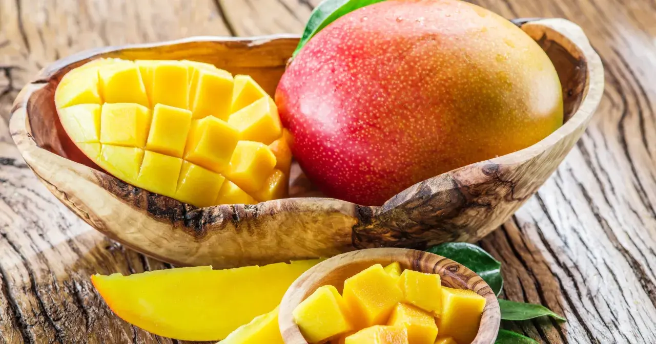 Dwa soczyste mango, jedno w całości, drugie pokrojone w kostkę, w drewnianych miseczkach. Idealne na zdrową przekąskę, mango kcal to ok. 60 na 100g.