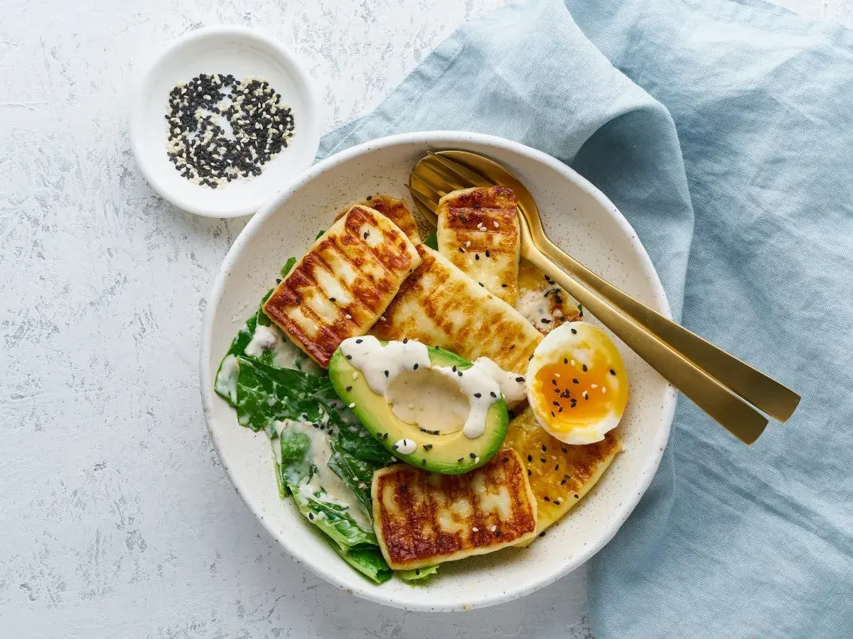 Sałatka z grillowanym serem halloumi, awokado, jajkiem i zielonymi liśćmi, posypana sezamem.
